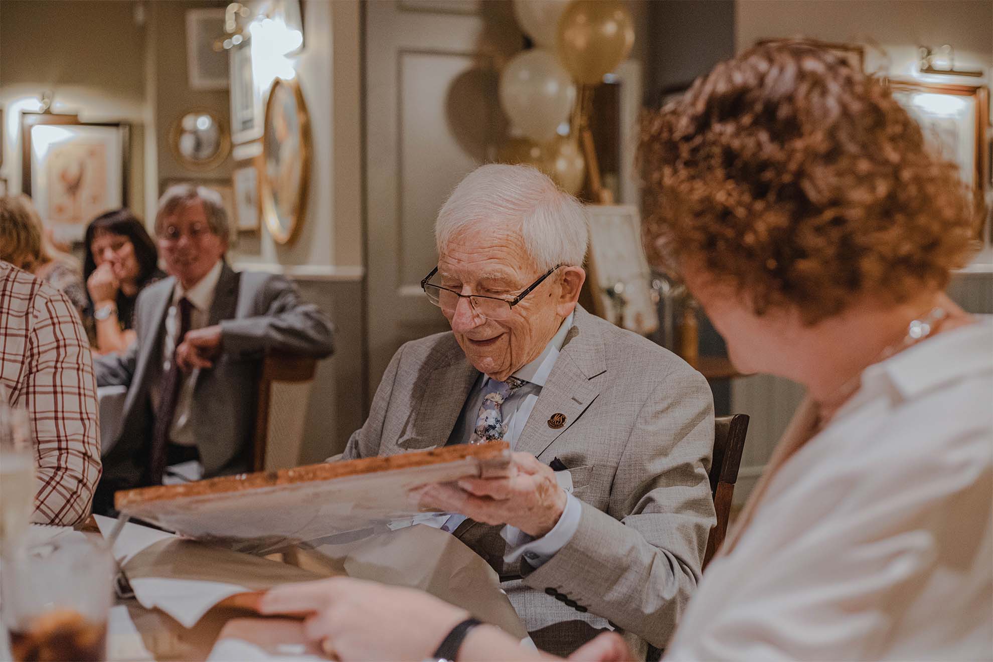 older man in a suit sat down opening a gift at a celebration